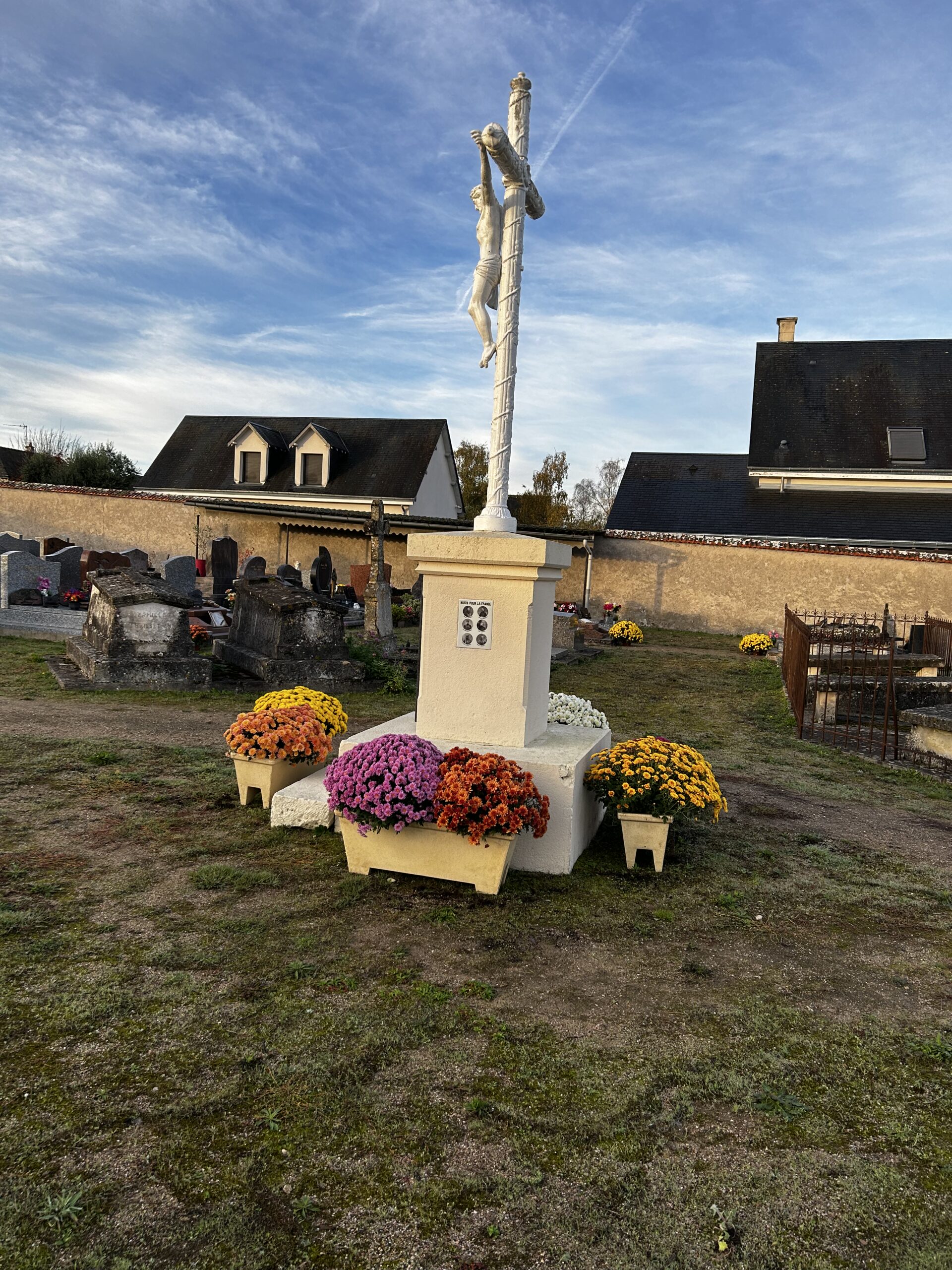 Christ en croix sur un calvaire de pierre se détachant sur un ciel bleu, avec des chrysanthèmes multicolores à son pied.