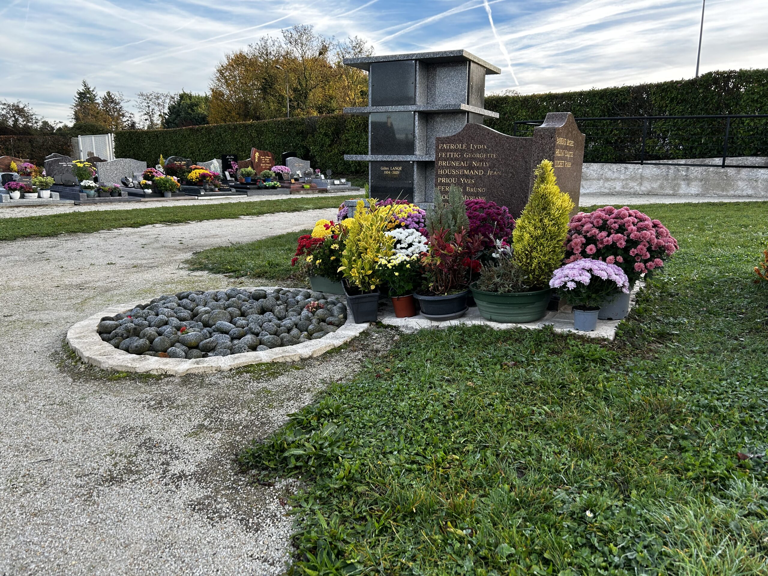 Tombe fleurie de chrysanthèmes blancs, jaunes et roses, accompagnée de plaques commémoratives et de petits objets de souvenir.